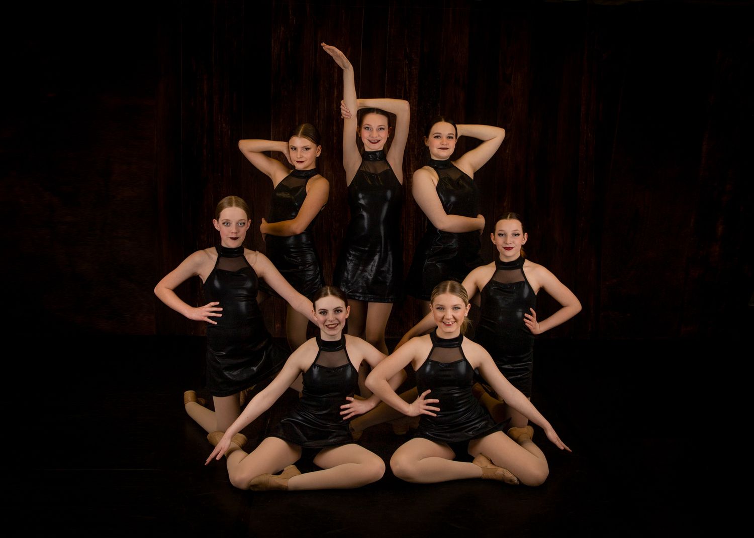 Dance group strikes dramatic pose in black outfits against dark background.