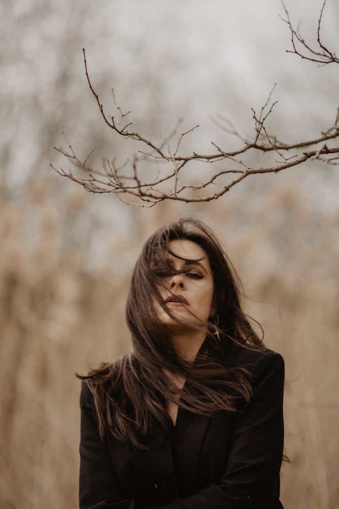 Dramatic portrait with wind-blown hair against bare branches in muted tones.