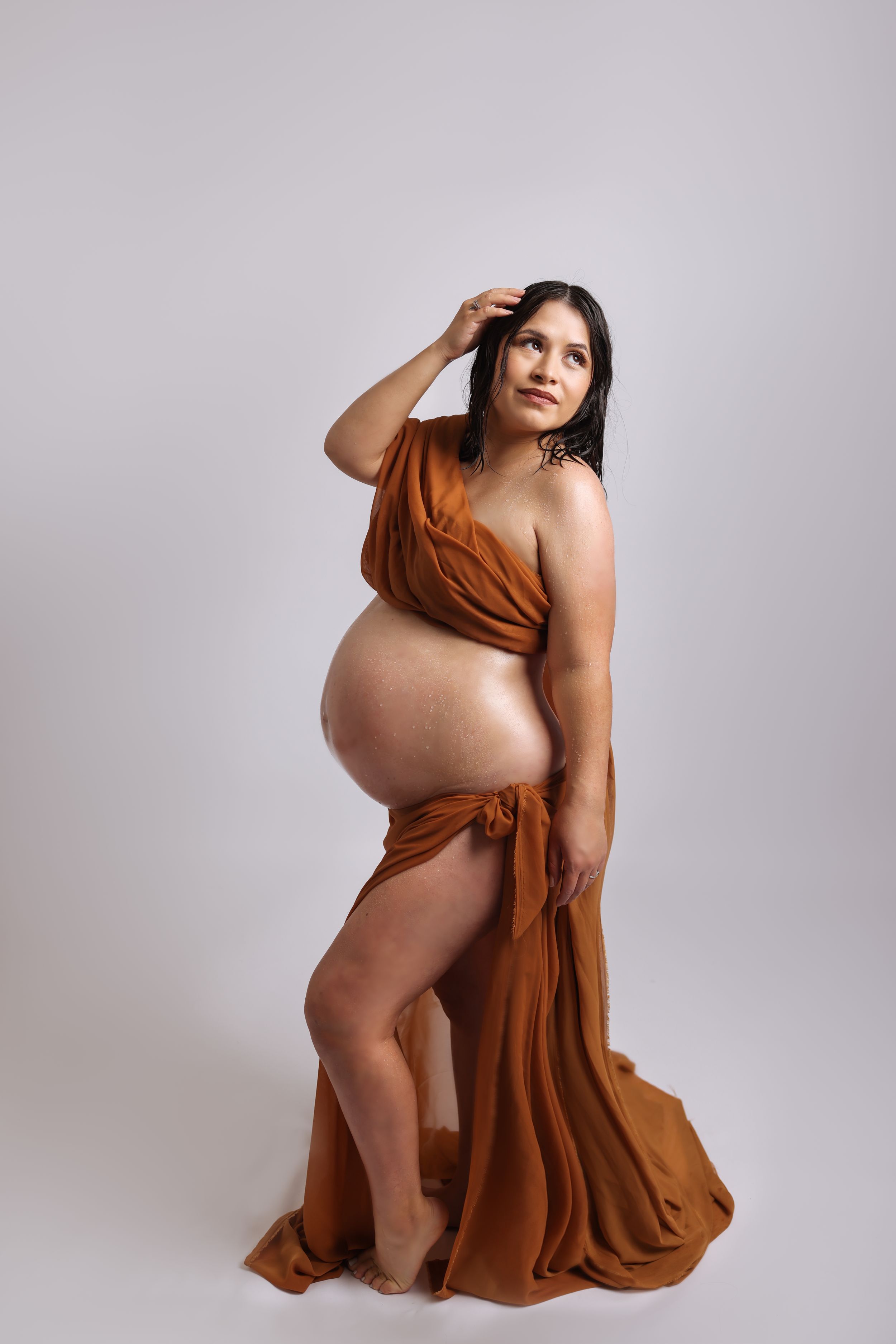 Artistic maternity portrait in draped rust-colored fabric against light background.