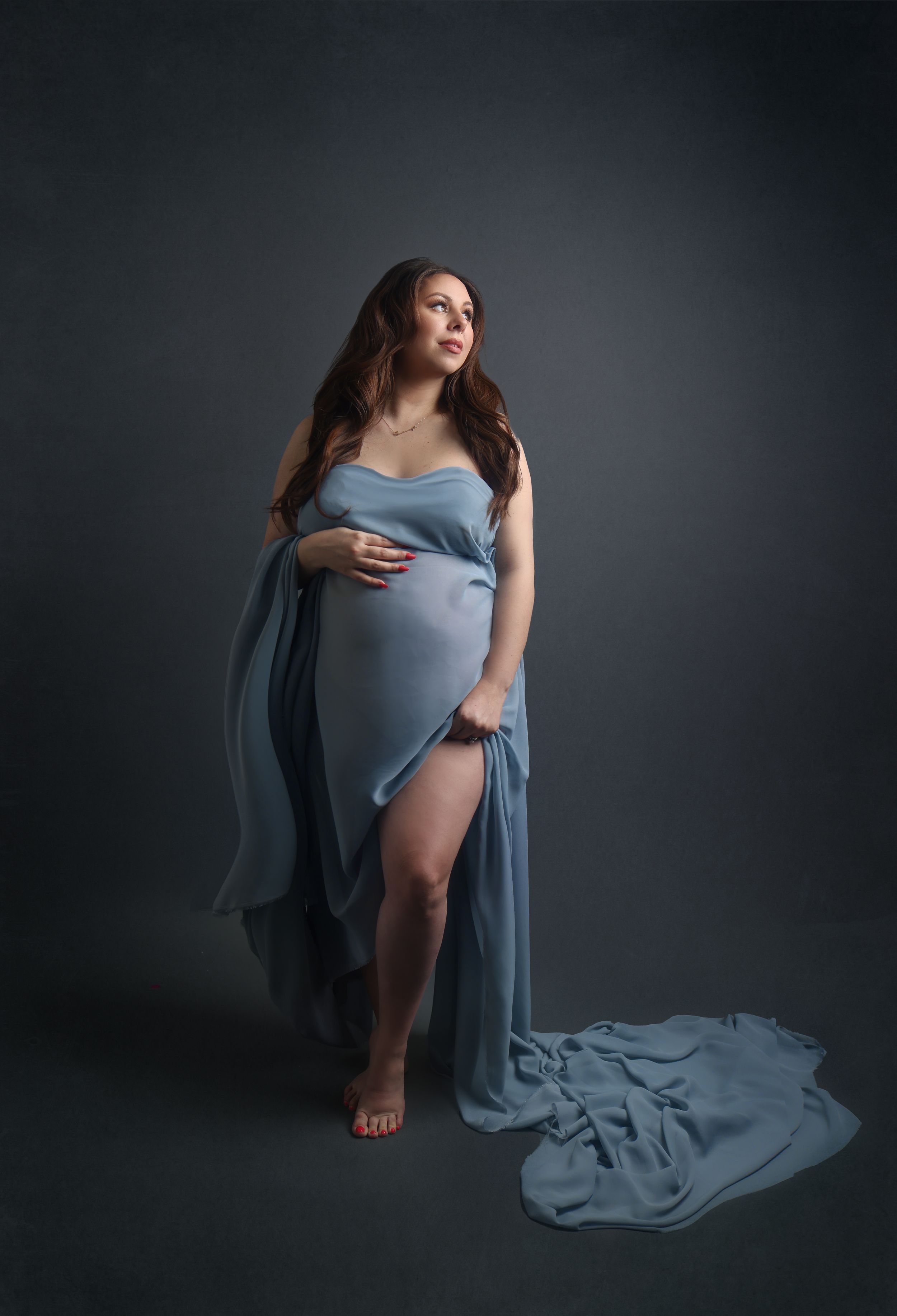 Ethereal maternity portrait in flowing blue dress against dark backdrop.