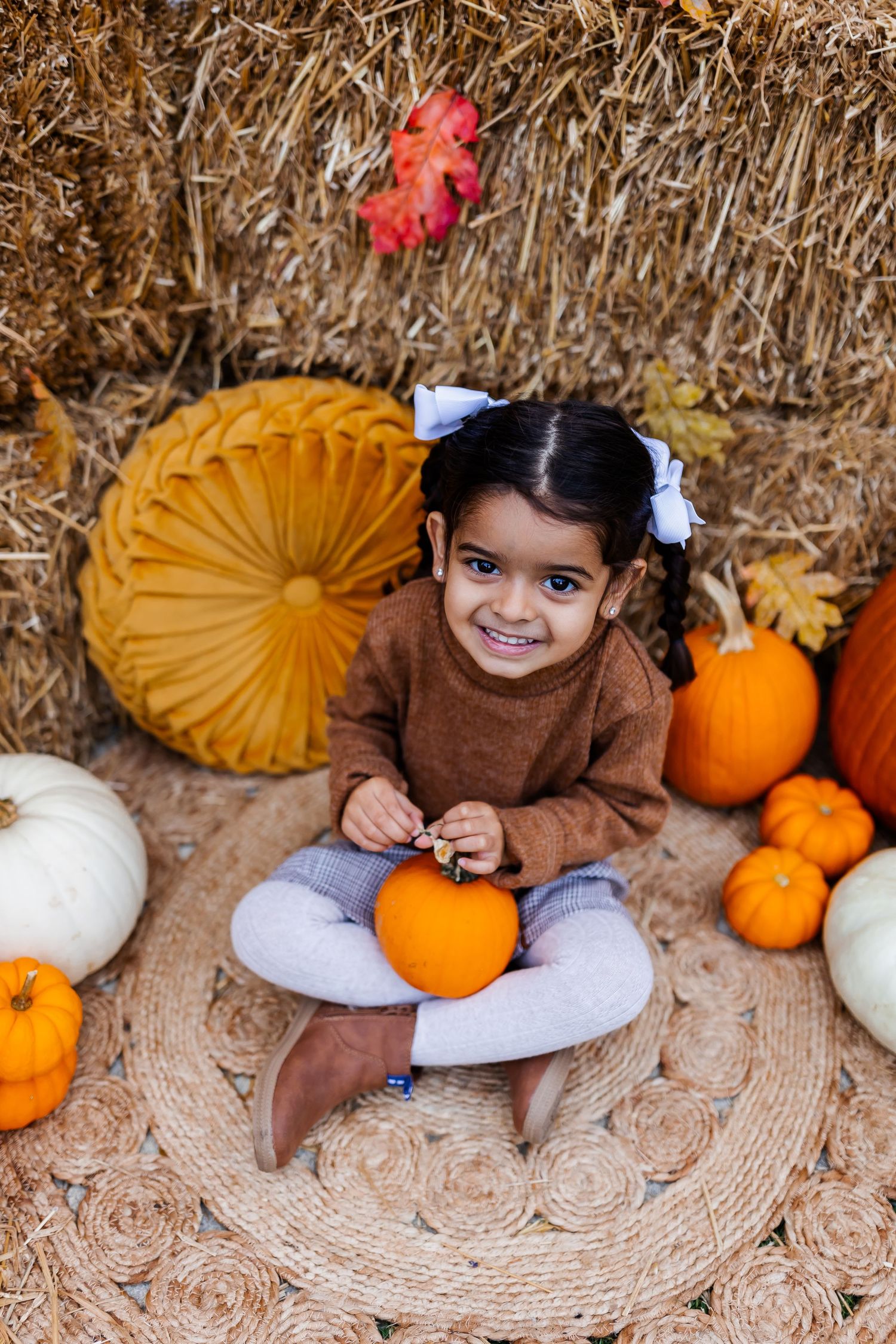 A child poses among decorative pumpkins and hay bales in an autumn themed photoshoot setting.