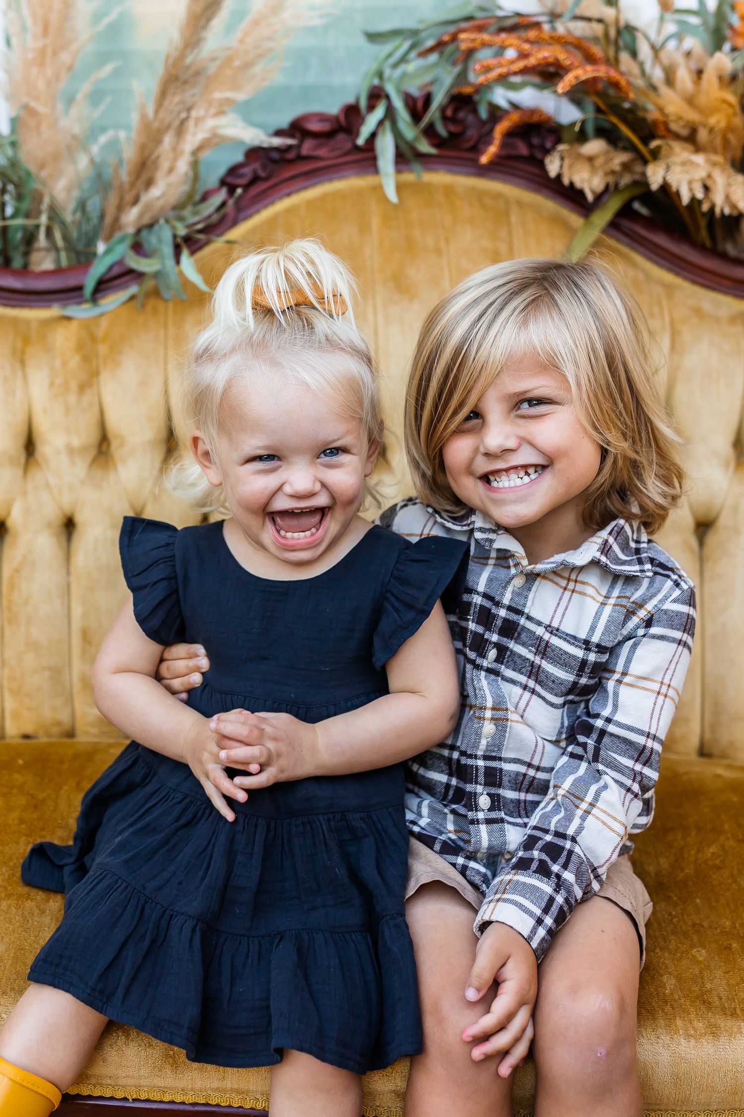 Two children in black and plaid outfits laugh together on a yellow couch.