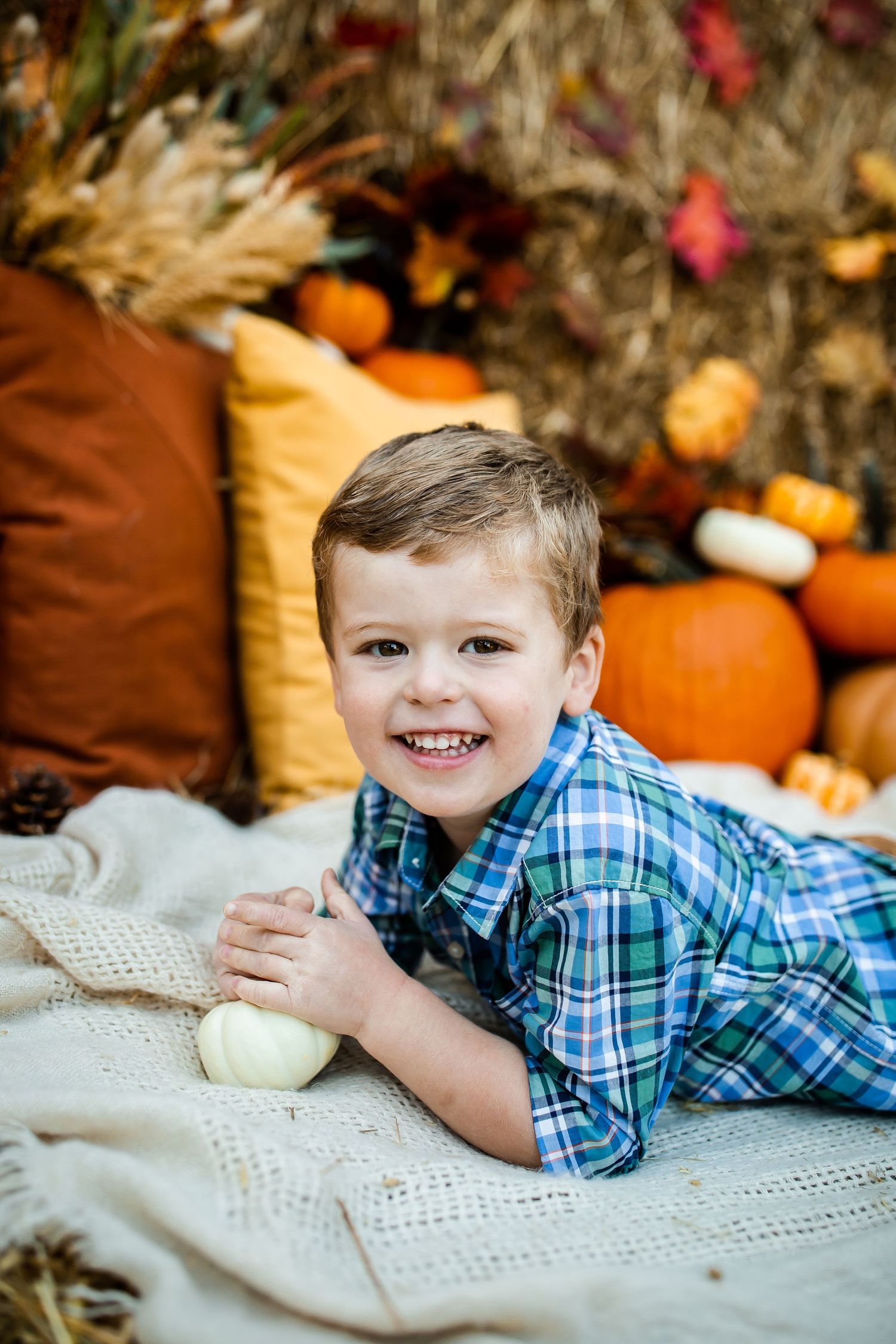 A plaid-wearing subject lies on a blanket among fall decorations including pumpkins and autumn leaves.