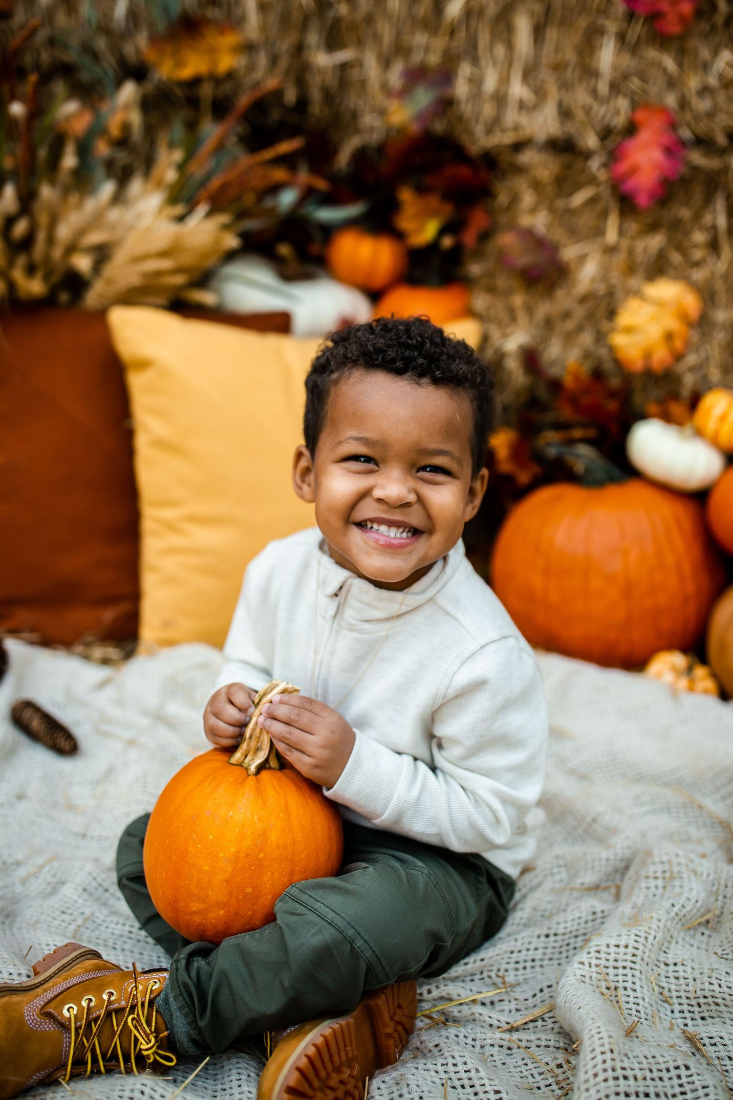 Small child in white sweater plays with orange pumpkins on textured blanket with fall backdrop.