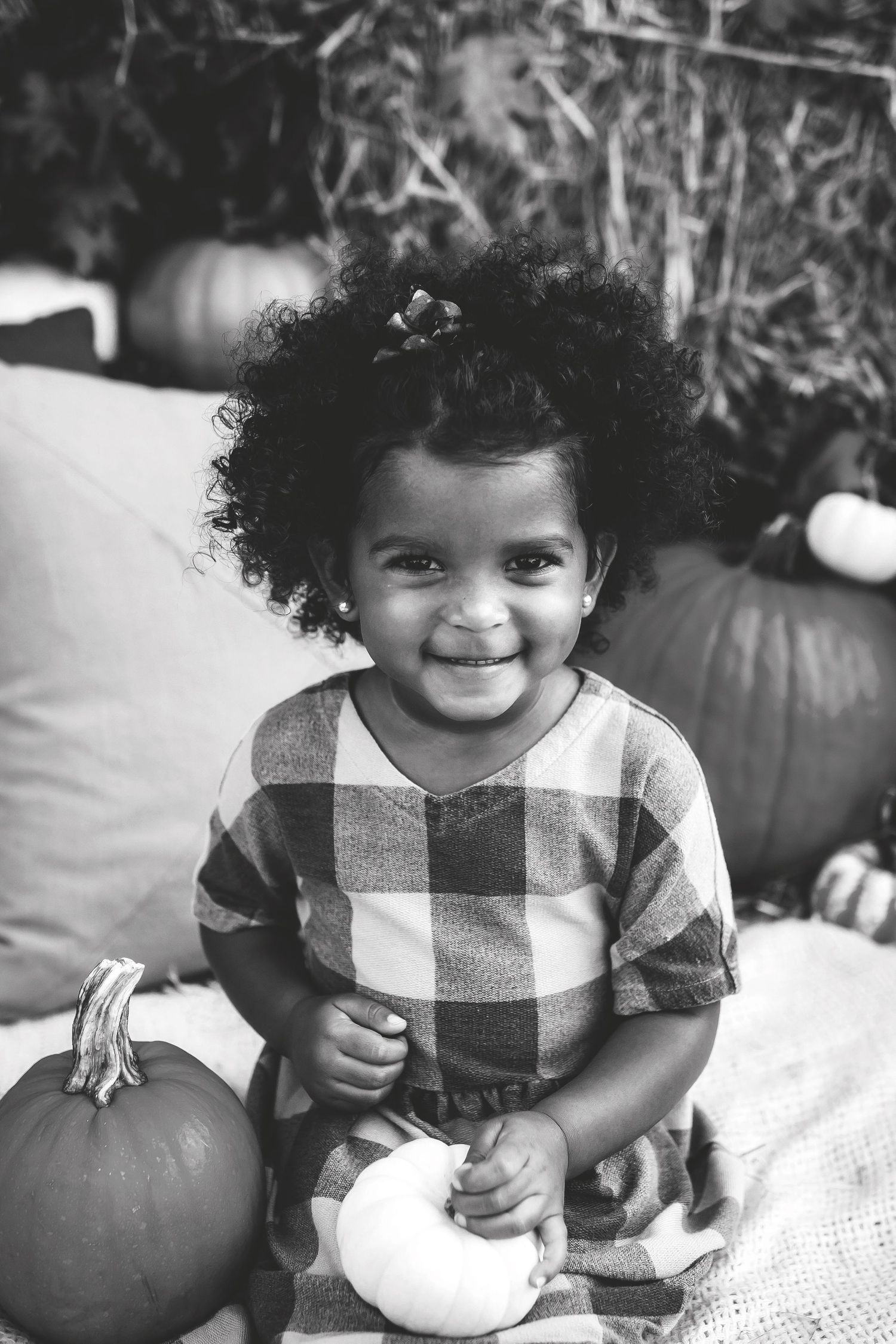 Black and white portrait of a toddler in a checkered shirt sitting with white and orange pumpkins.