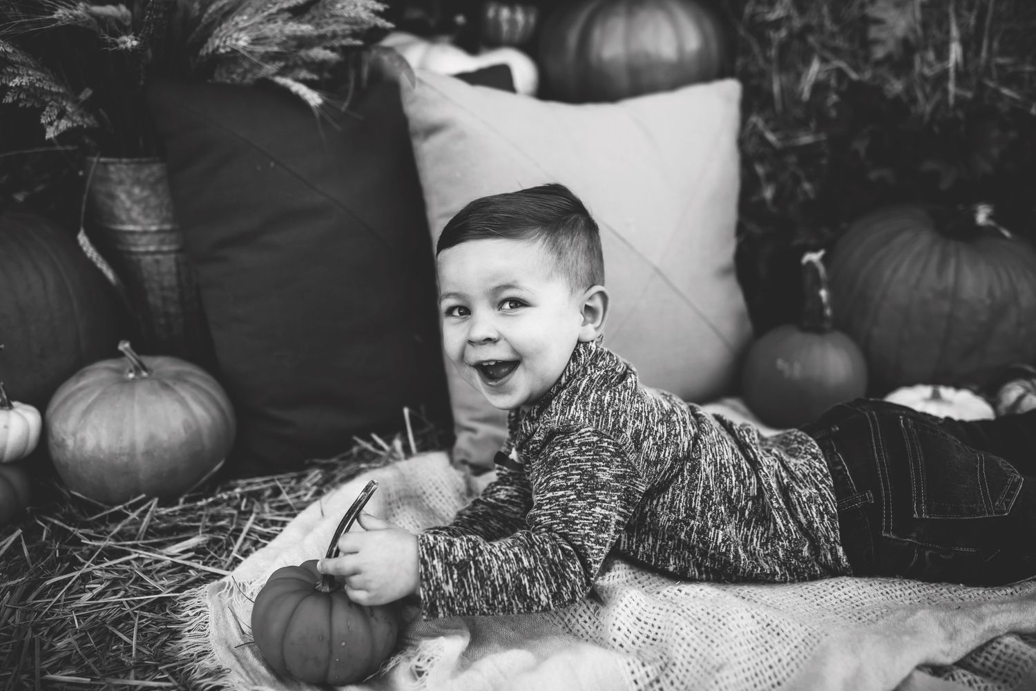 Black and white photo of a child lying on hay with pumpkins during autumn.