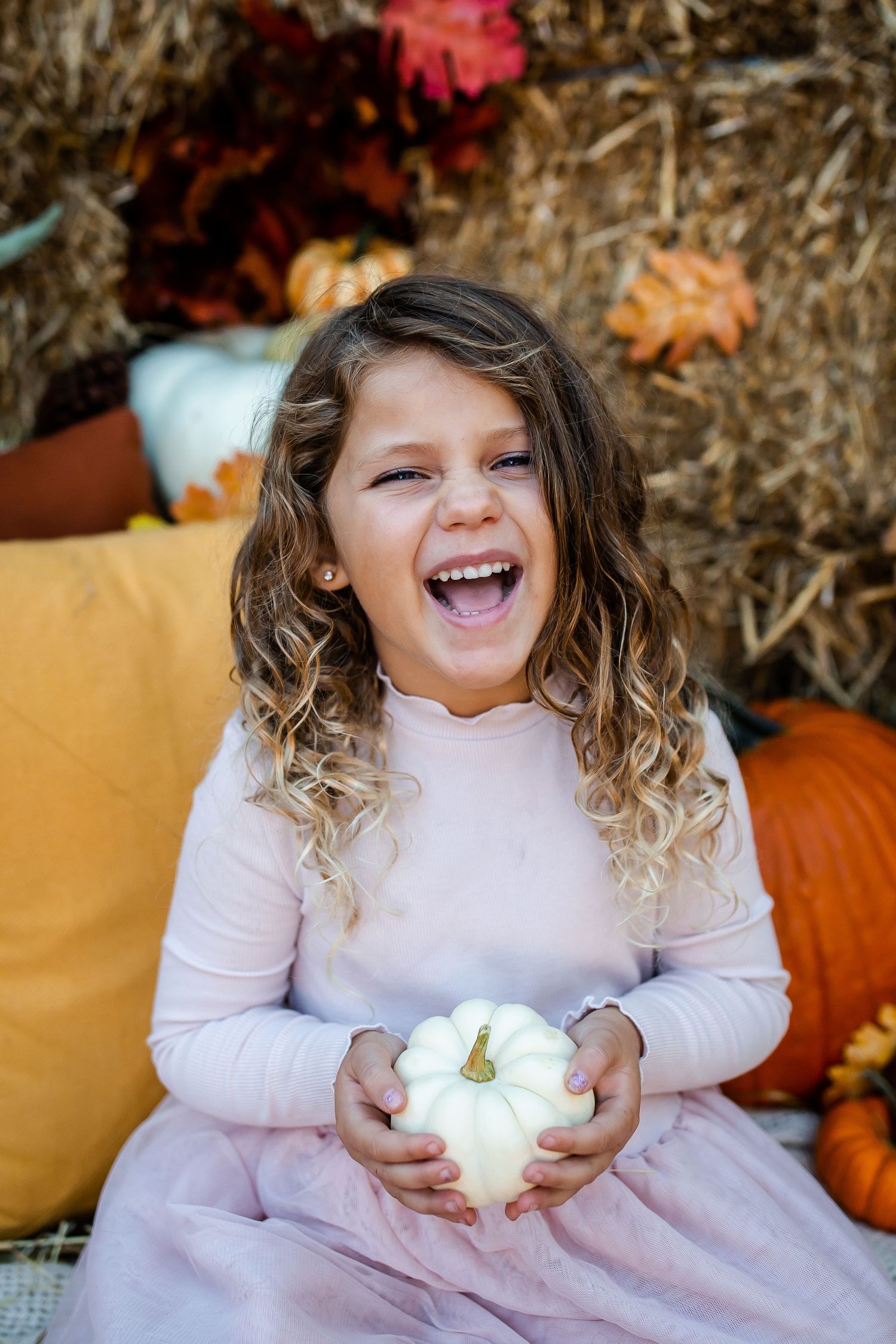 Child in pink dress holds white pumpkin against autumn backdrop with hay and orange pumpkins.
