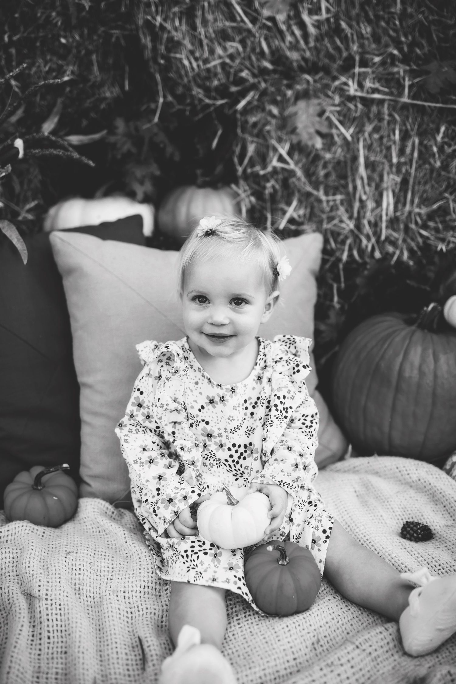 Baby dressed in polka dot dress poses with white pumpkins on crocheted blanket.