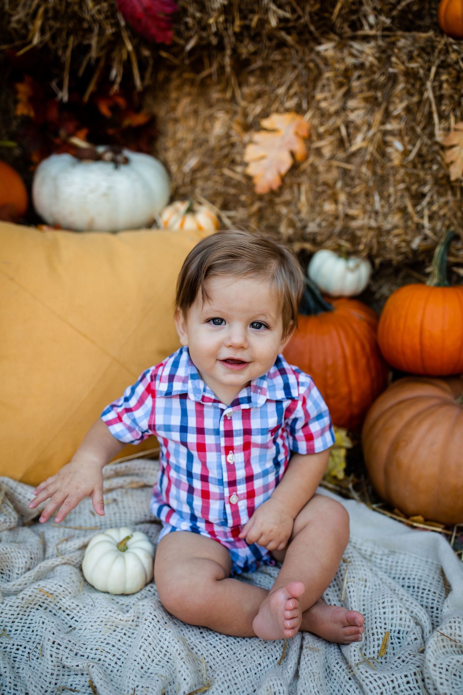 Baby in plaid shirt poses with white and orange pumpkins on lace blanket.