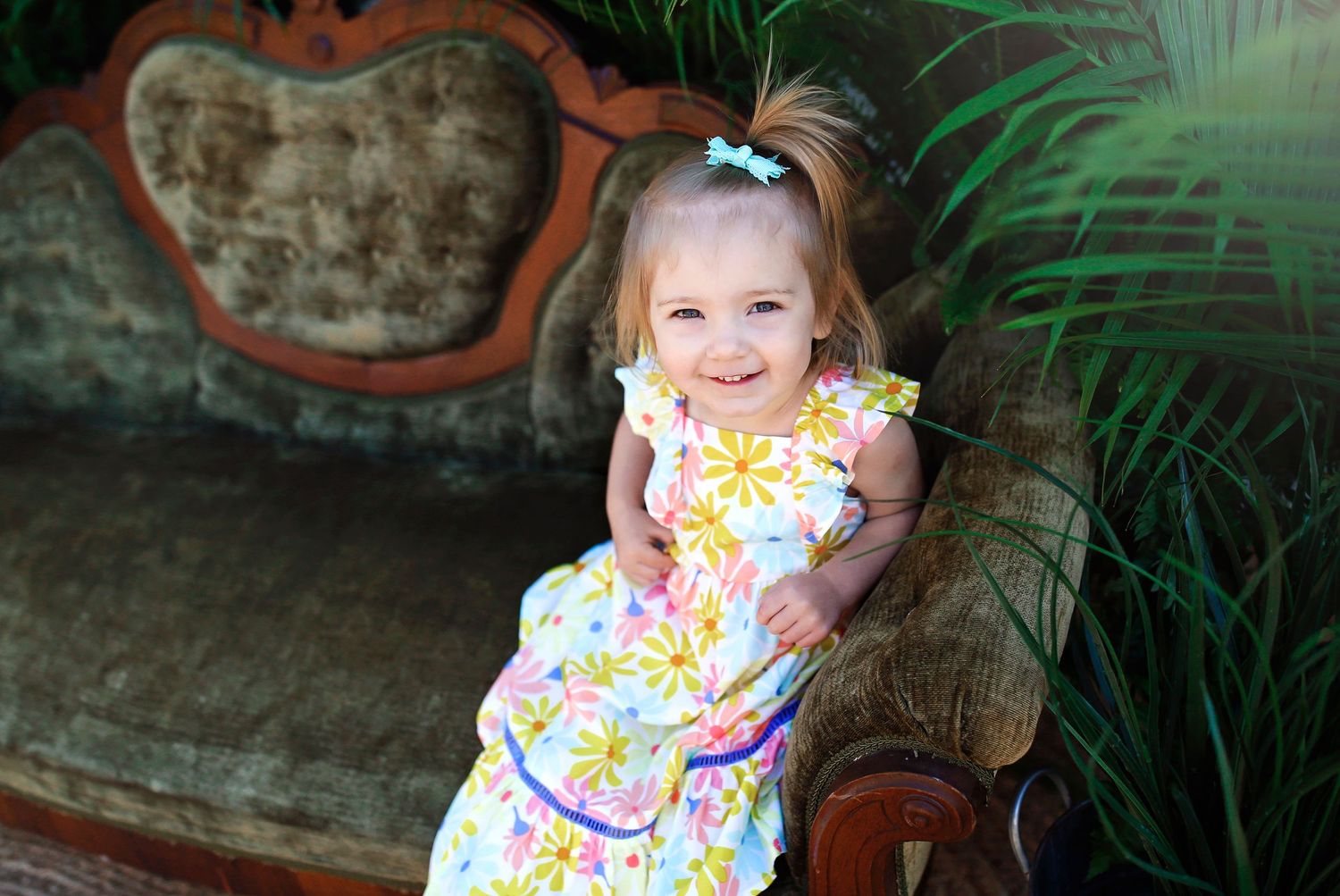 Child wearing yellow floral dress sitting on antique couch with tropical greenery.