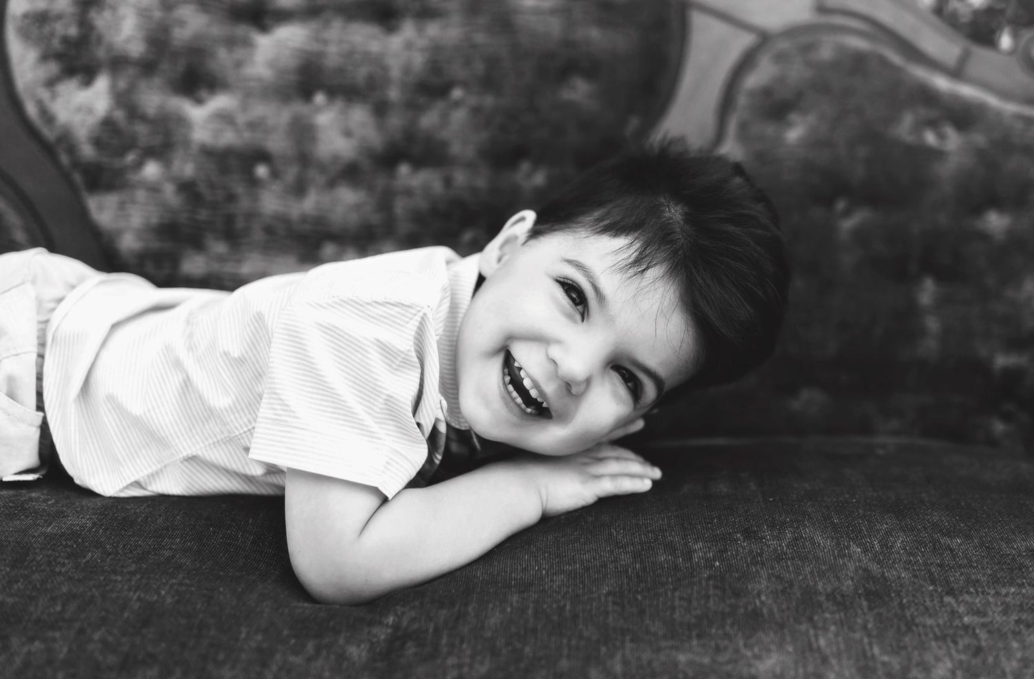 Black and white capture of child lying down on velvet couch with bright expression.