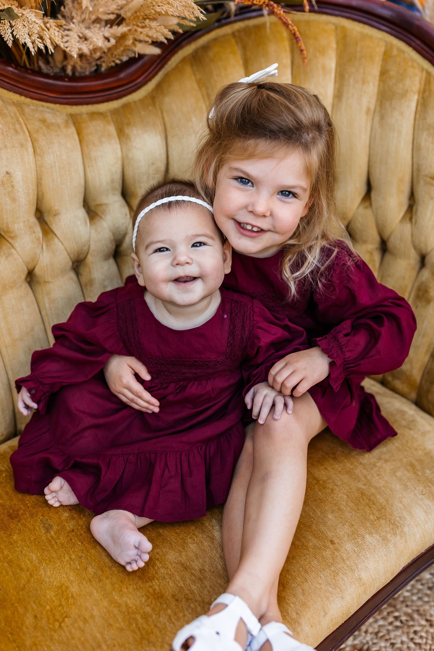 Two children in burgundy outfits share adorable moments on a tufted gold couch.