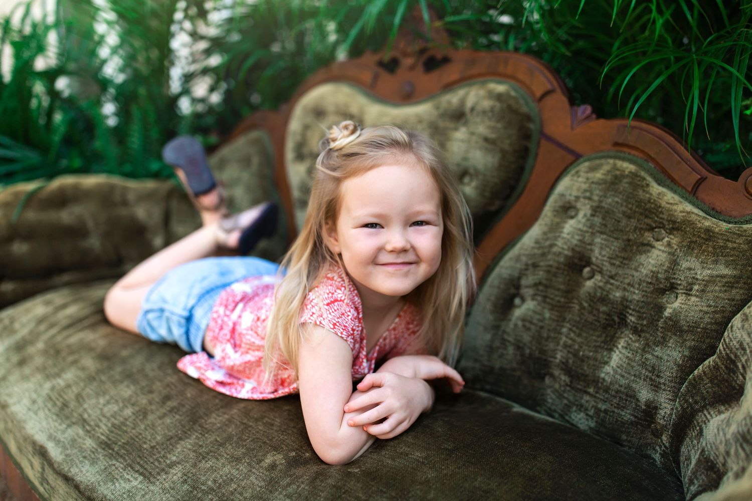 Child lounging on vintage green velvet sofa surrounded by tropical plants.