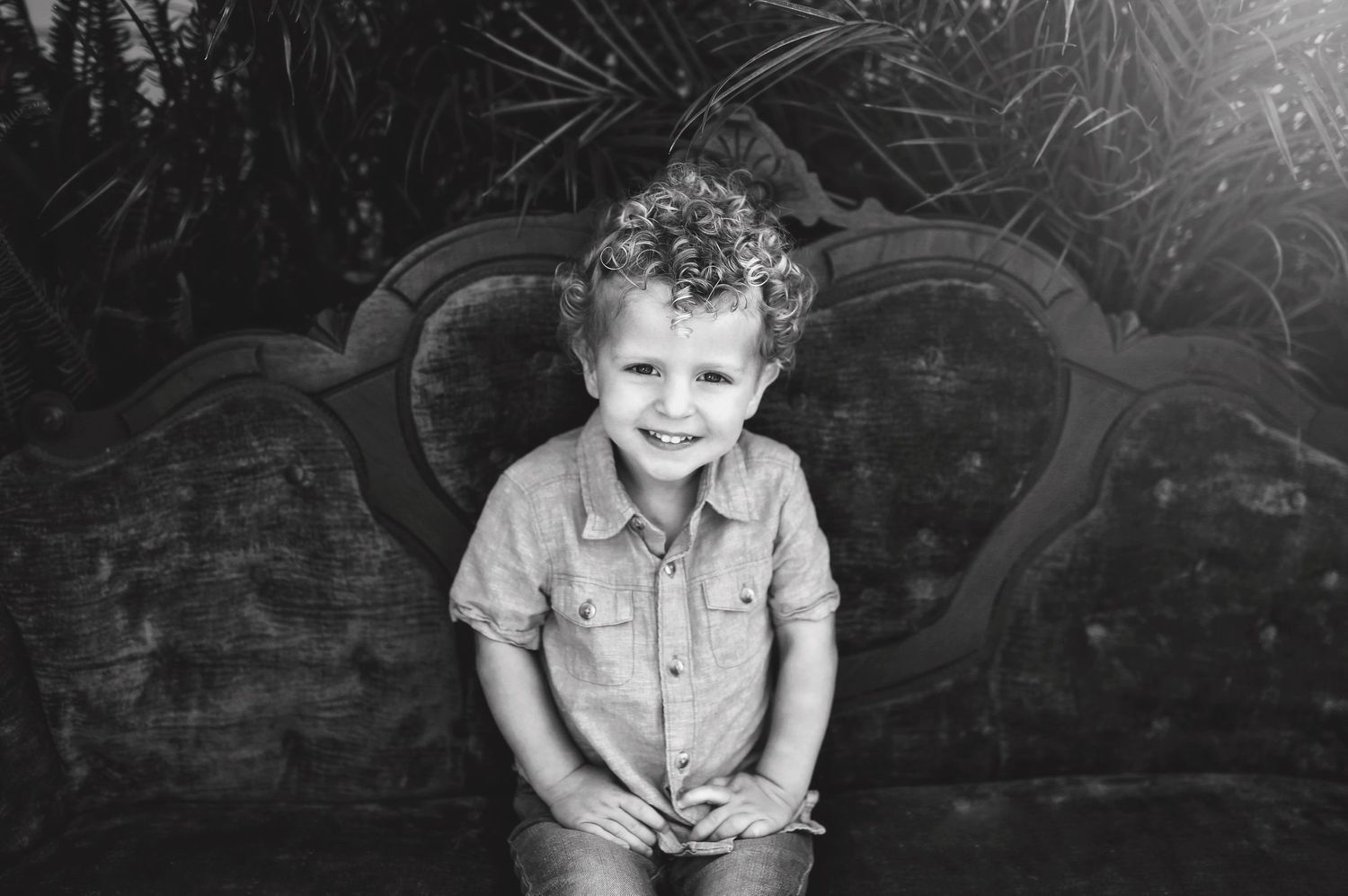 Black and white portrait of child standing against vintage sofa backdrop.