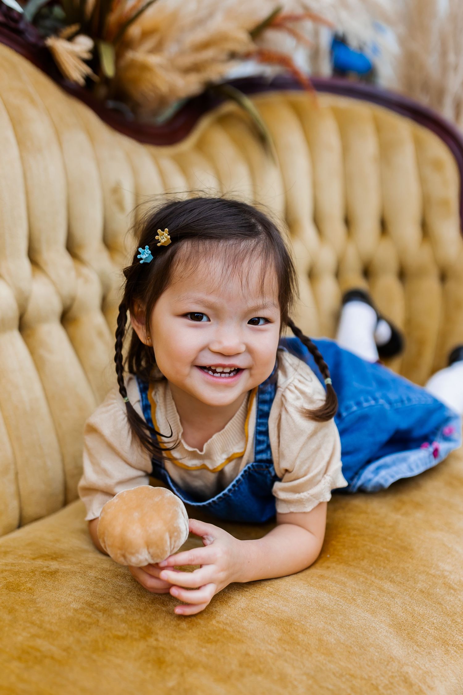 A child in denim plays on a yellow tufted couch during a photo session.