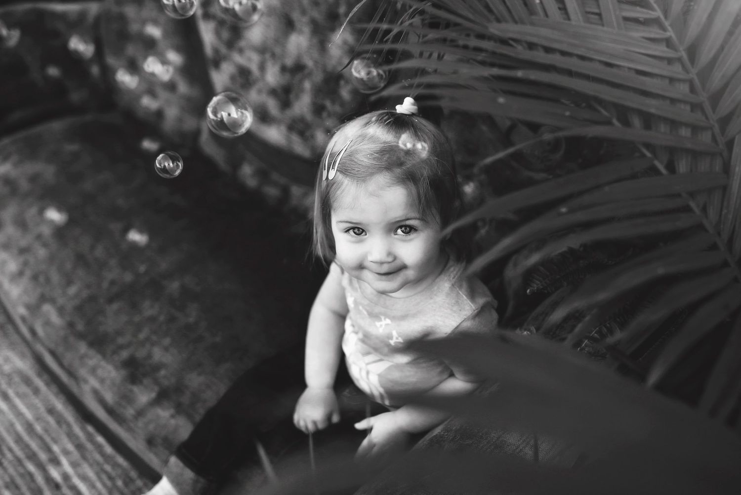 Black and white photo of child on vintage furniture with soap bubbles and palm fronds.