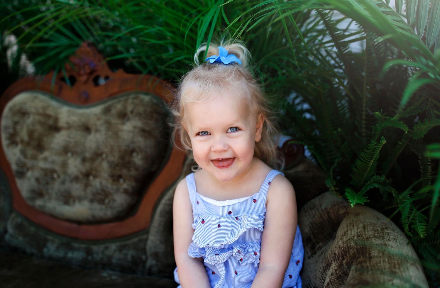 Child in blue dress with hair bows sitting on antique sofa among tropical plants.