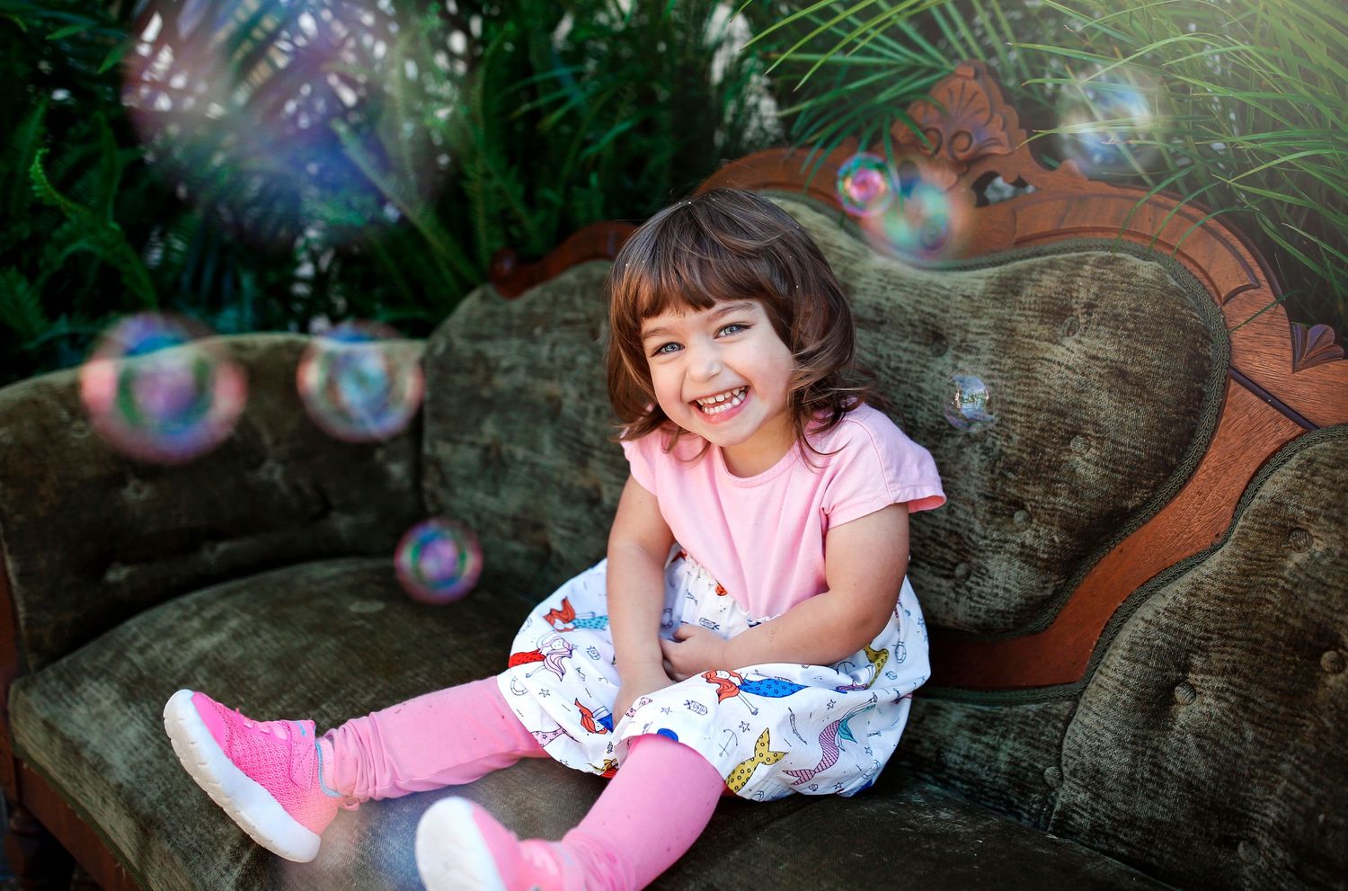 Child in pink outfit sitting on vintage sofa with floating bubbles in background.