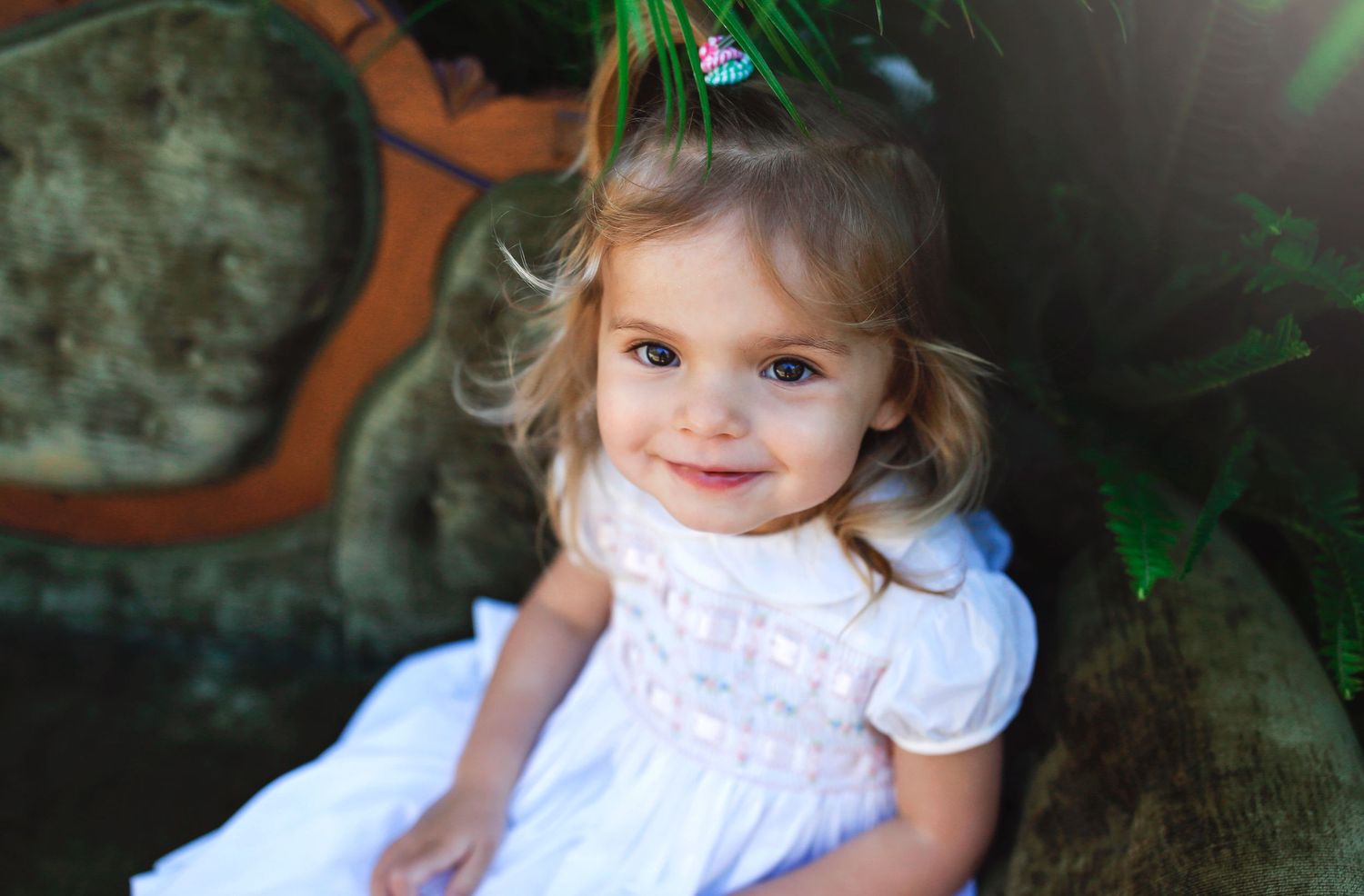 Young child in white dress sitting on vintage couch with greenery in background.