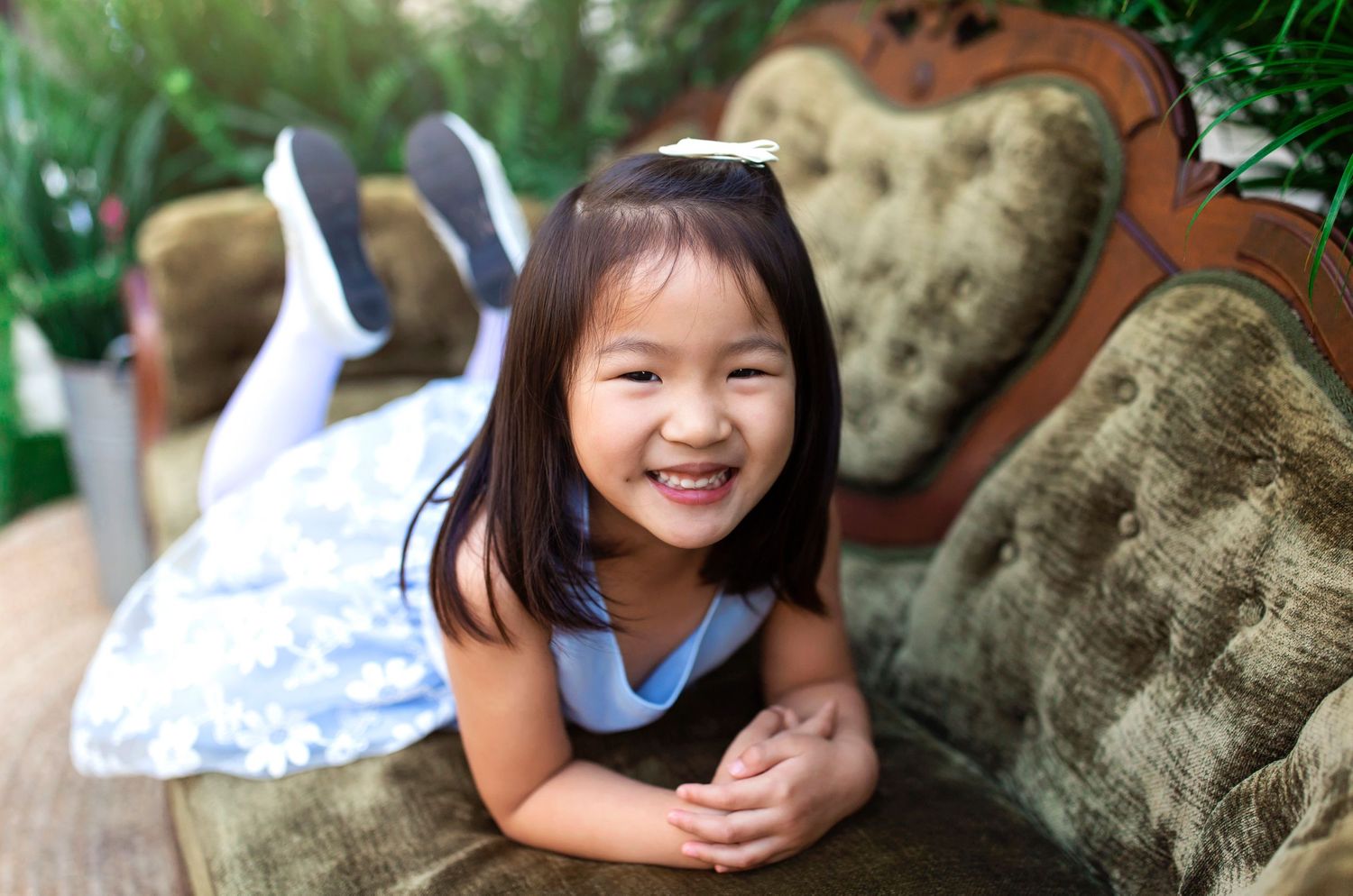 Child relaxing on antique velvet couch with tropical foliage backdrop.