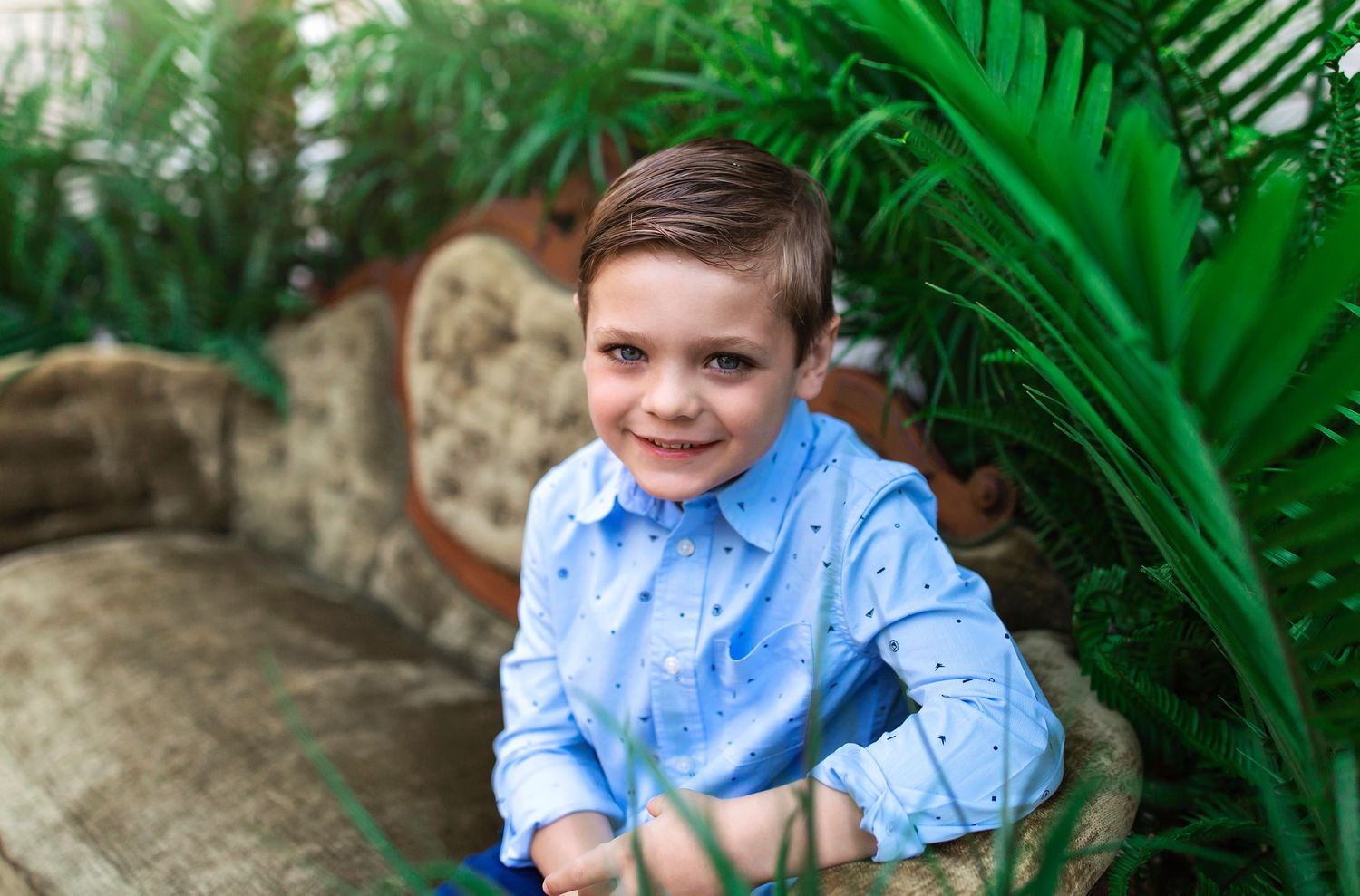 Young person in light blue shirt poses against tropical greenery backdrop.