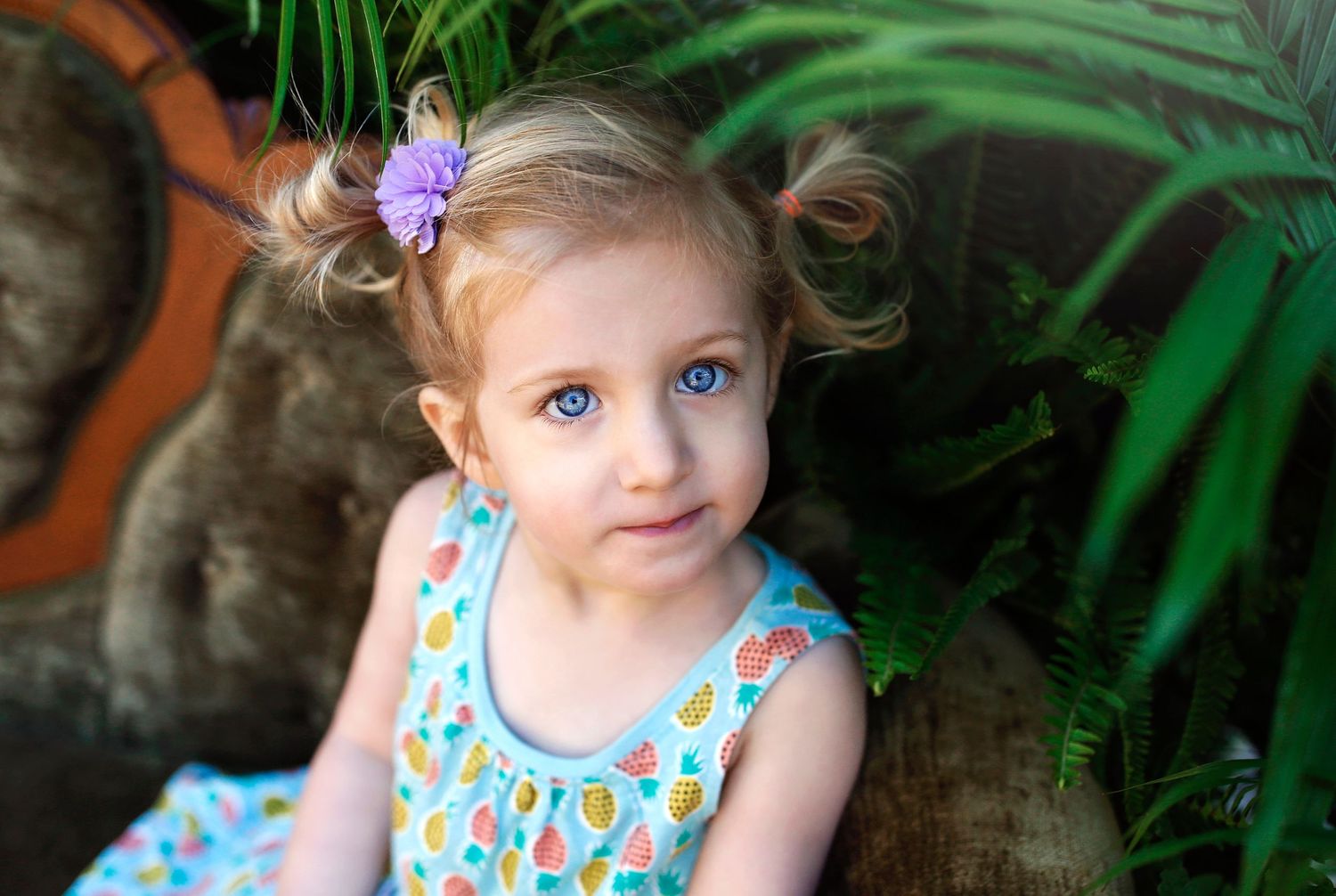 Child with purple flower hair accessory sitting against tropical plant backdrop.
