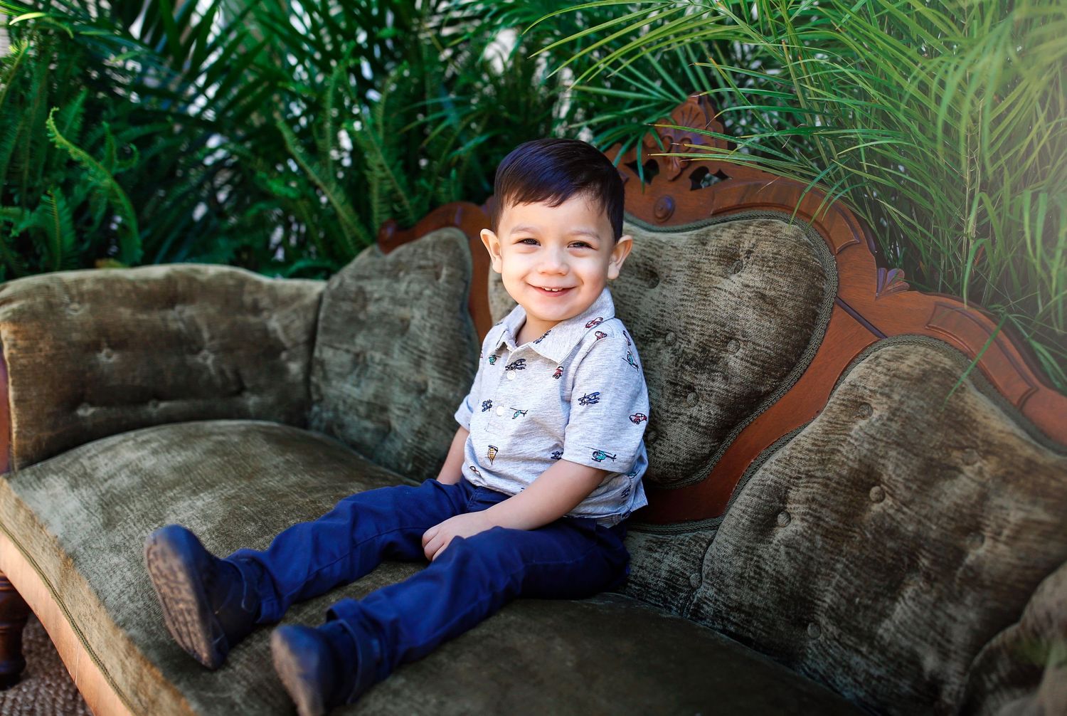 Child in button-down shirt sitting on antique velvet sofa surrounded by tropical plants.