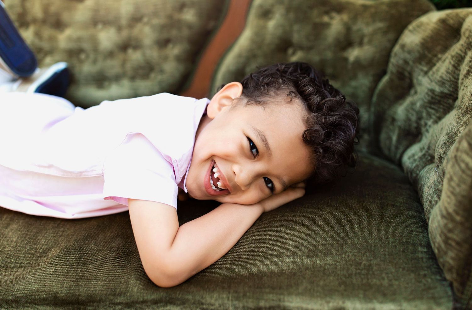 Child lying playfully on green velvet sofa with bright smile.