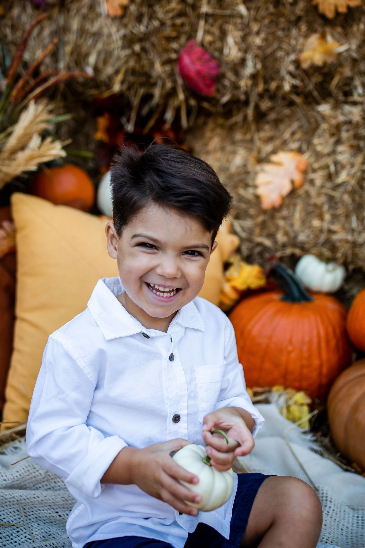 A subject in white shirt poses cheerfully with mini pumpkins against an autumn-themed backdrop.