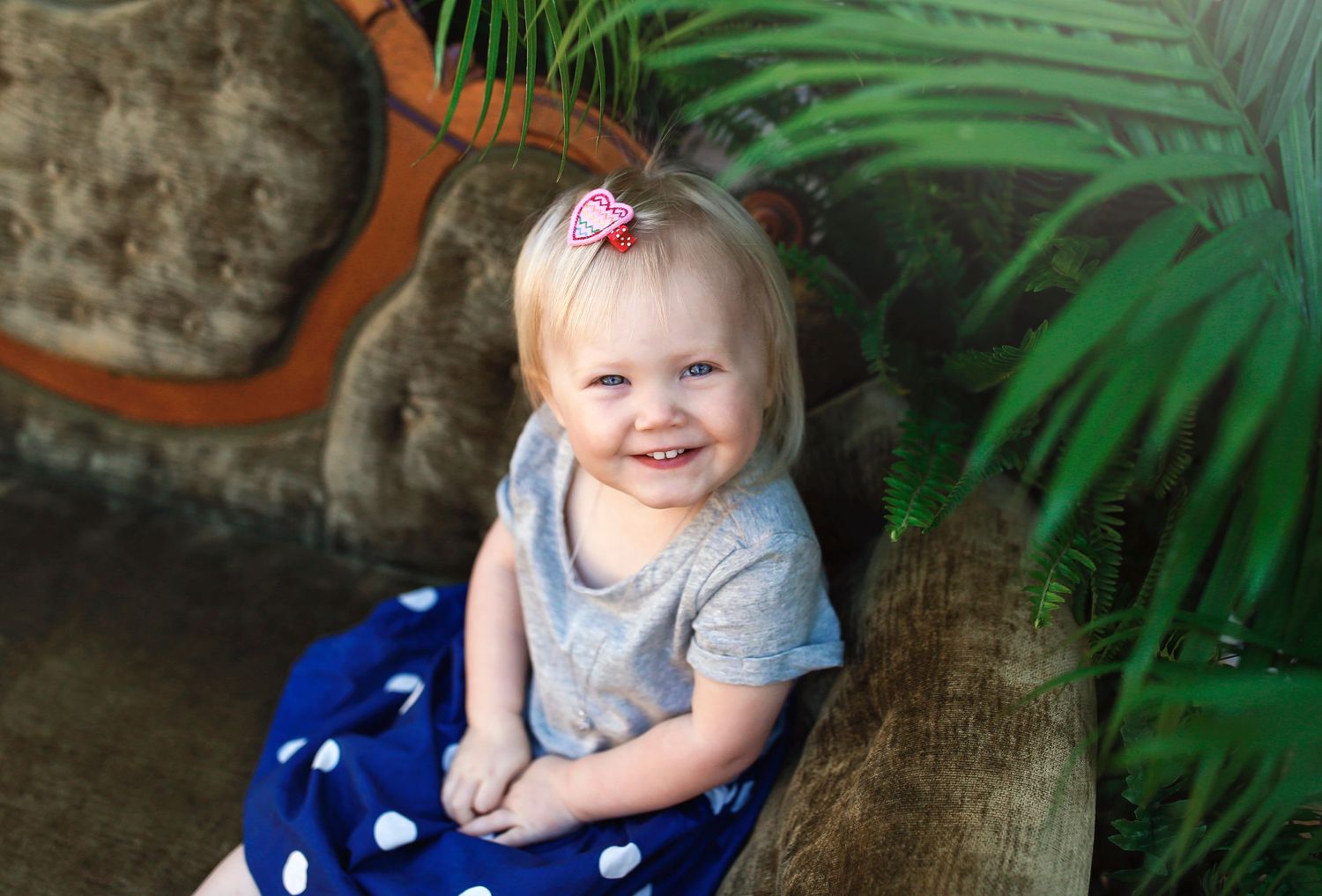 Child in grey shirt and polka dot outfit sitting on antique couch with palm leaves behind.