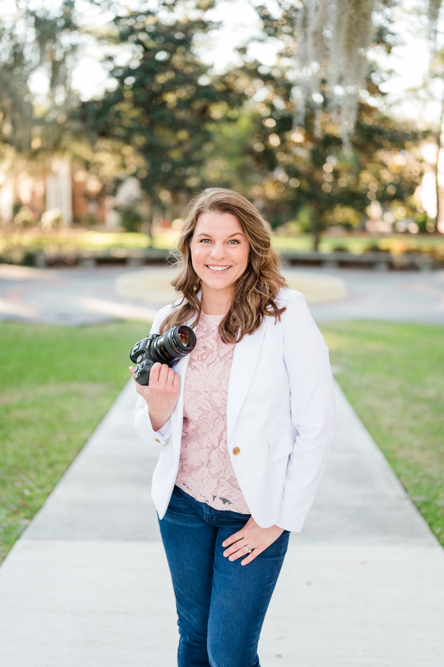 Professional photographer wearing a white blazer, pink blouse and jeans poses with camera outdoors in natural lighting.