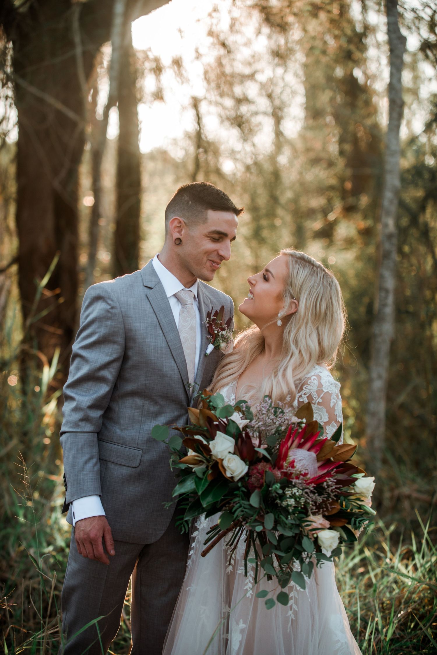 A wedding couple shares an intimate moment in a sun-dappled forest with a vibrant red bouquet.