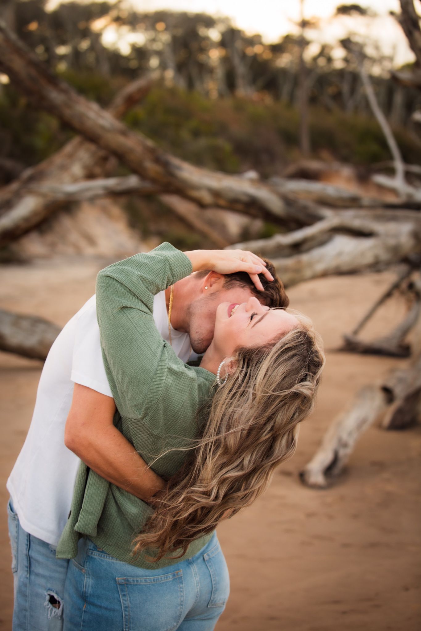 A romantic embrace captured at sunset on a beach with driftwood in the background.