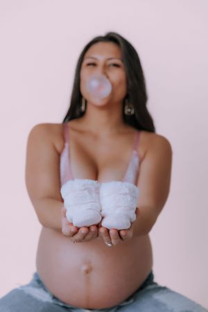 Maternity photo with white baby shoes placed on pregnant belly against pink background.