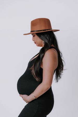 A pregnant woman in a black tank top and brown hat poses against a white background.