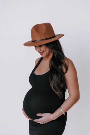 Maternity portrait series with brown hat and black tank top against white background.