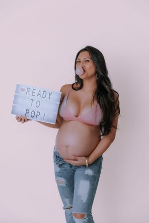 Pregnancy announcement photo holding 'Ready to Pop' sign in casual denim and pink top.