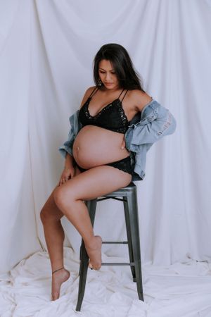 Studio maternity photos with woman in black lingerie and denim on chair.
