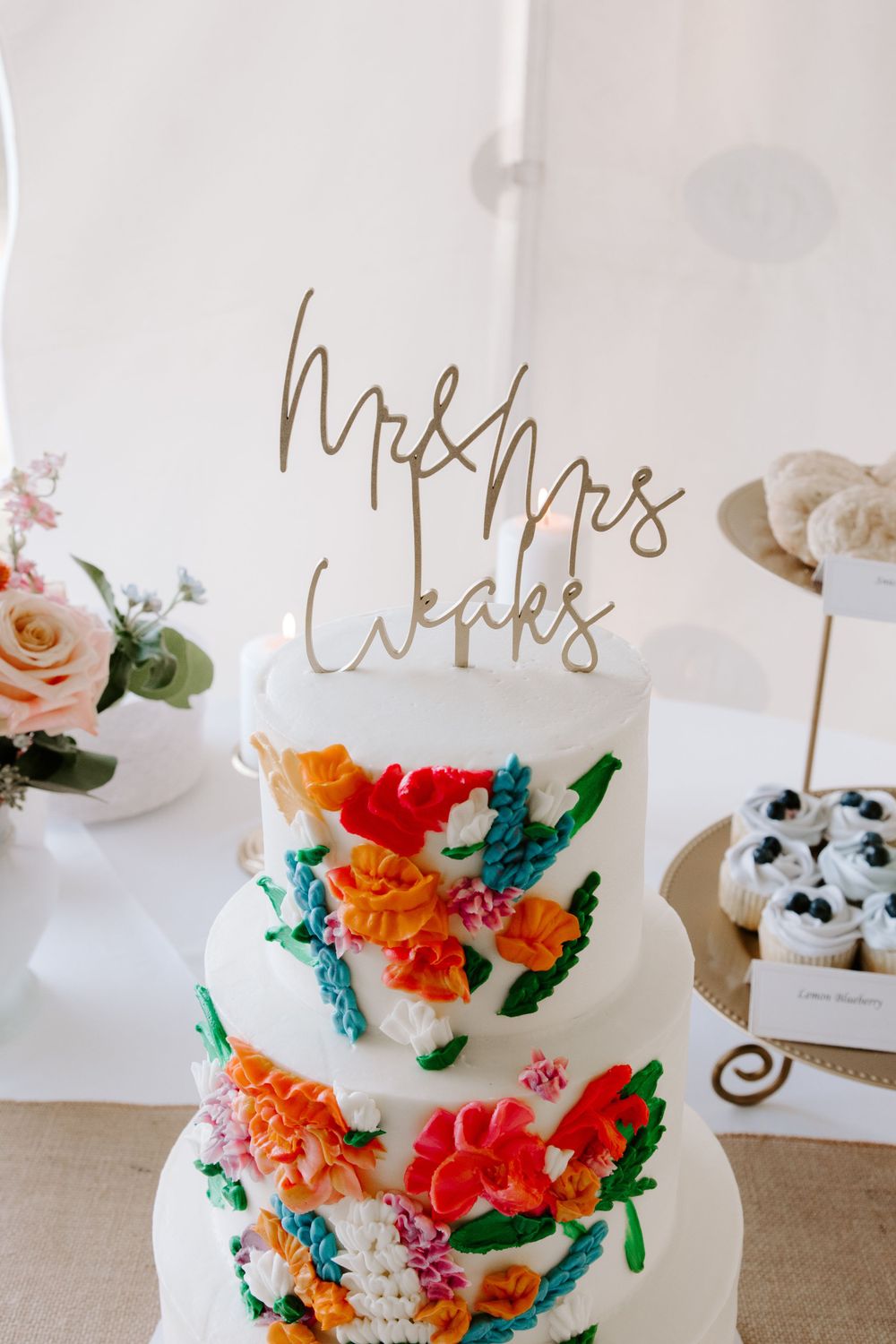 White wedding cake decorated with colorful flowers and gold 'Mr & Mrs Weeks' topper.