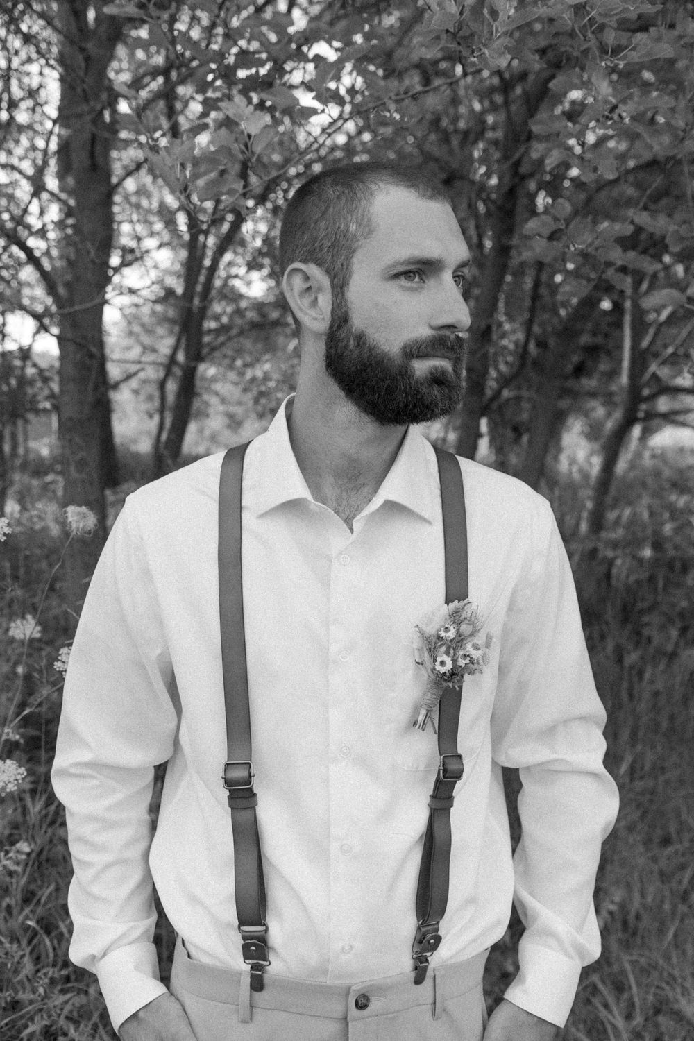 Groom in white shirt and suspenders poses in outdoor woodland setting.