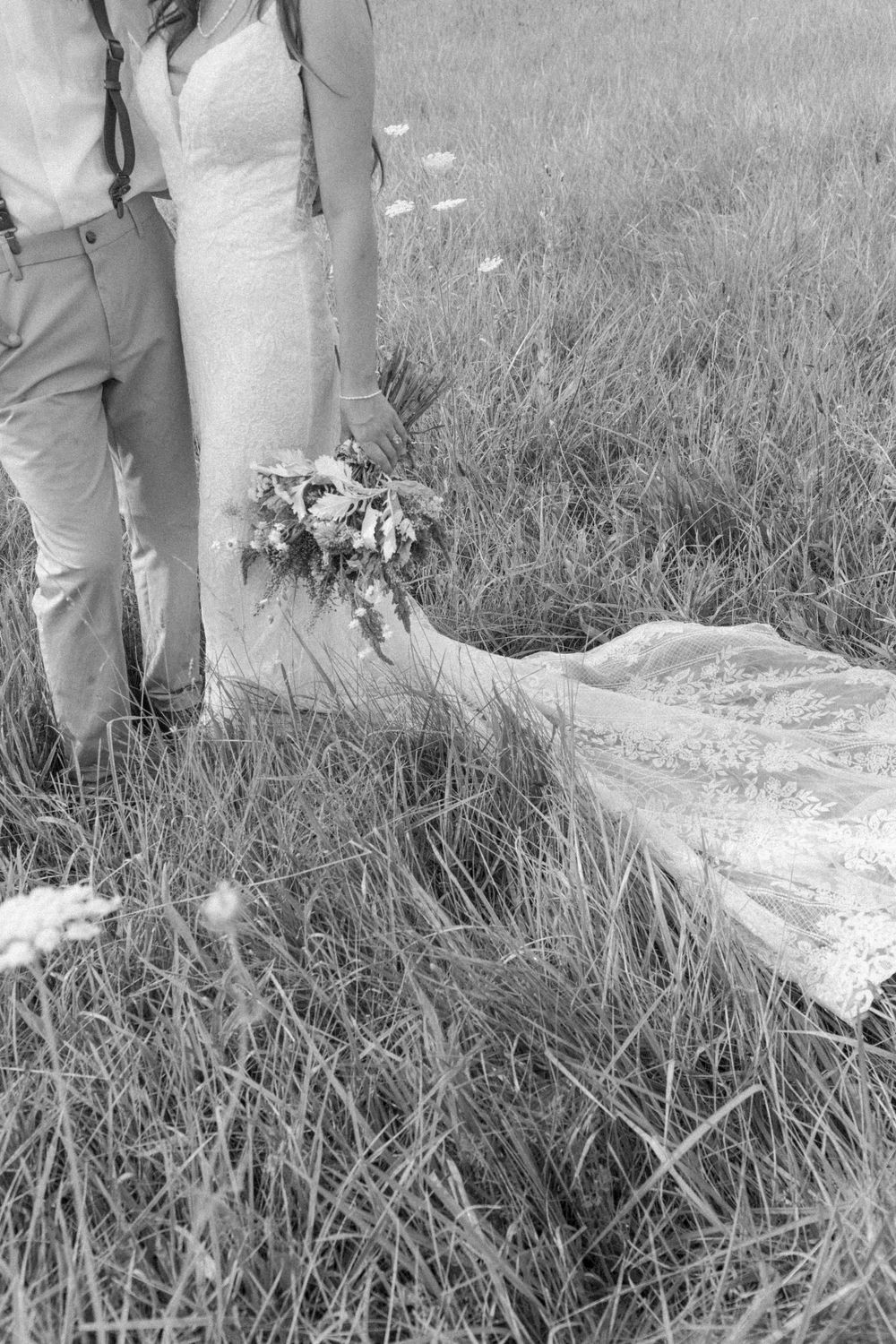 Black and white detail of wedding dress and bouquet trailing in tall grass.