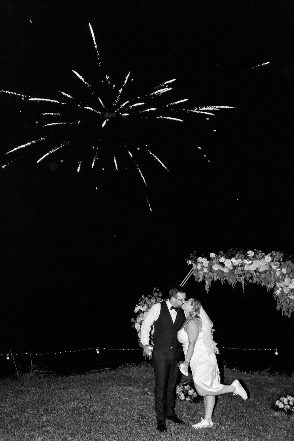 Romantic wedding moment under floral arch with fireworks display in night sky.