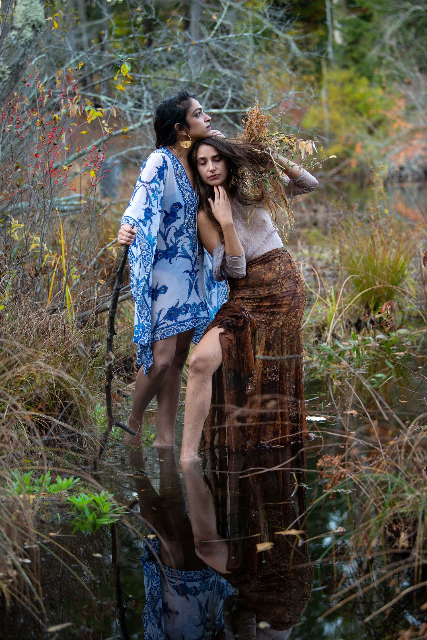 Two figures in flowing dresses pose artistically by pond with autumn foliage reflections.