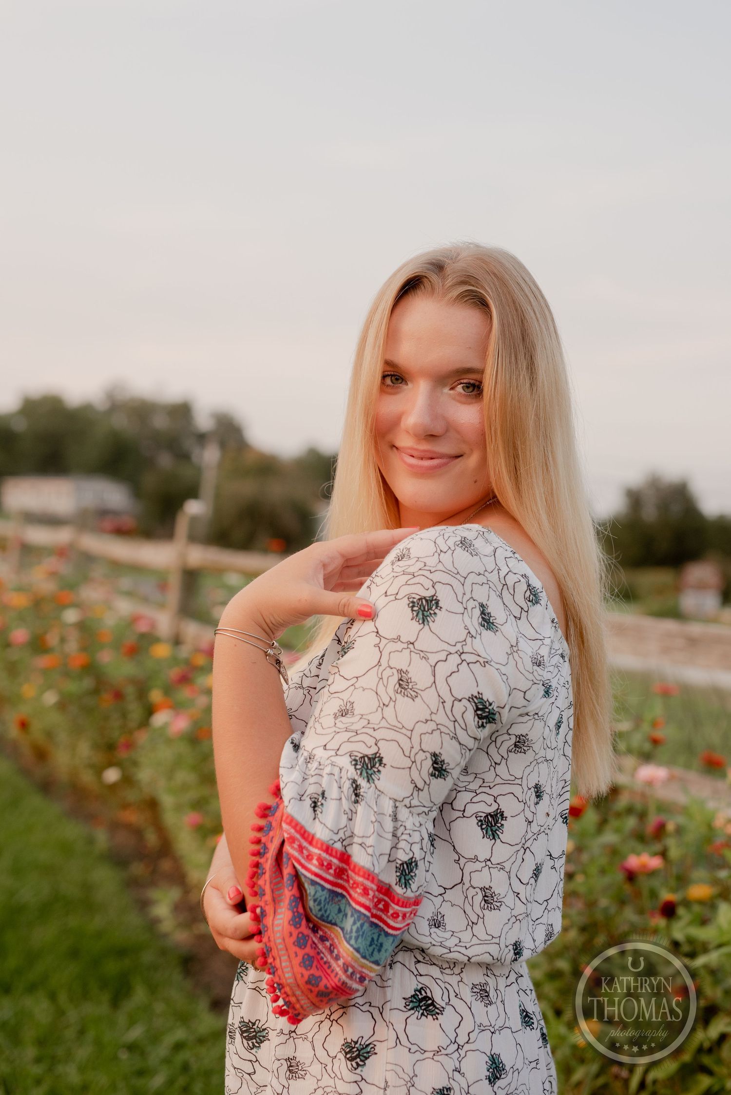 Woman in black and white floral dress poses near colorful flower garden at dusk.