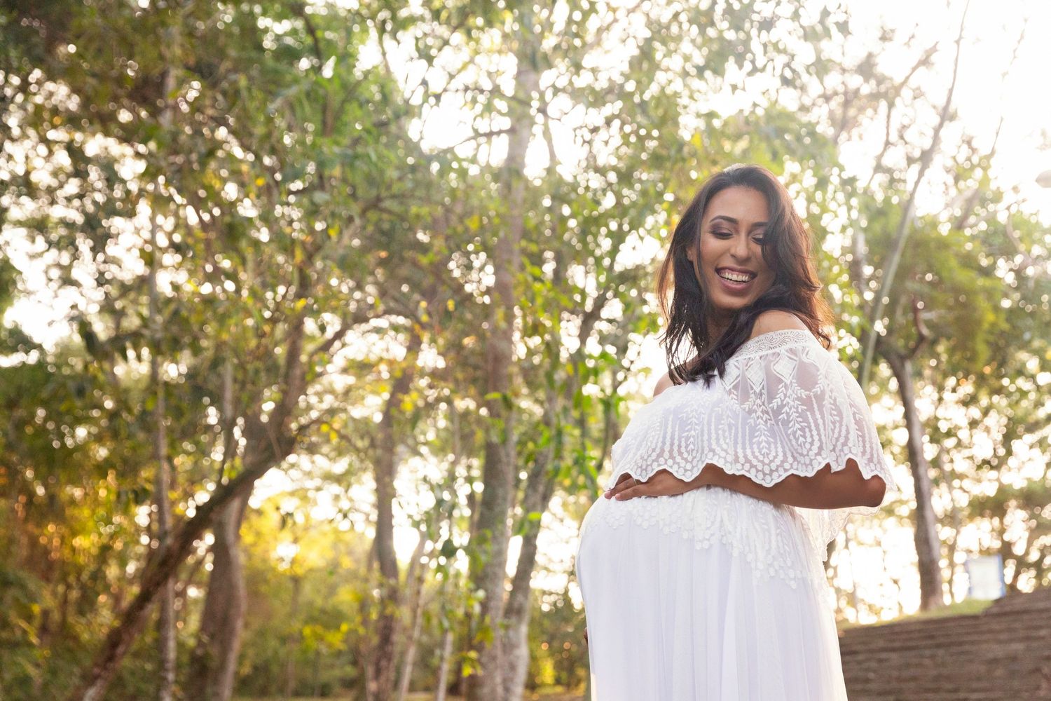 Woman in white lace dress standing in sunlit wooded area.