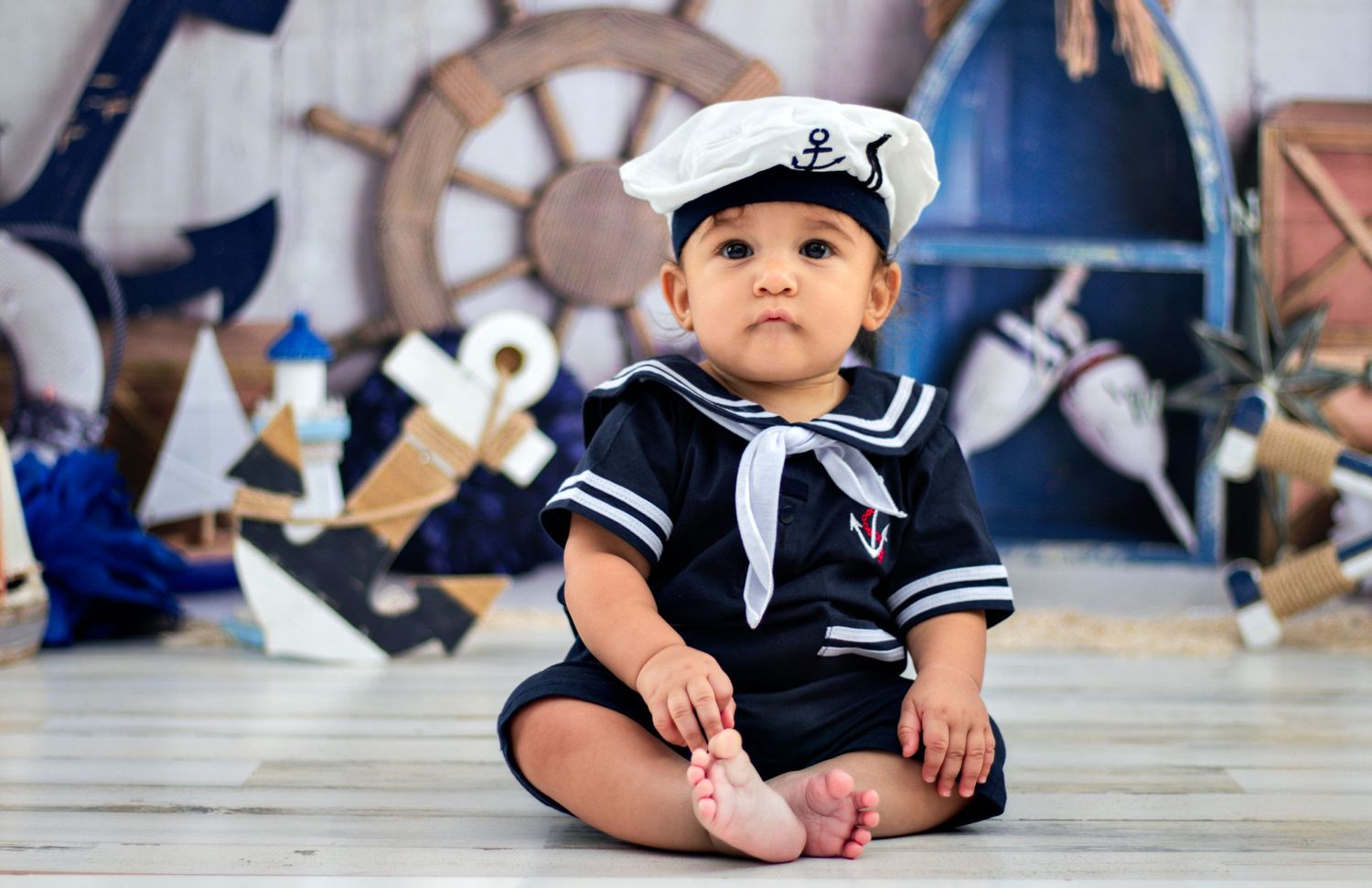 A baby wearing a white and navy sailor outfit with matching hat sits on a wooden floor with nautical decorations.