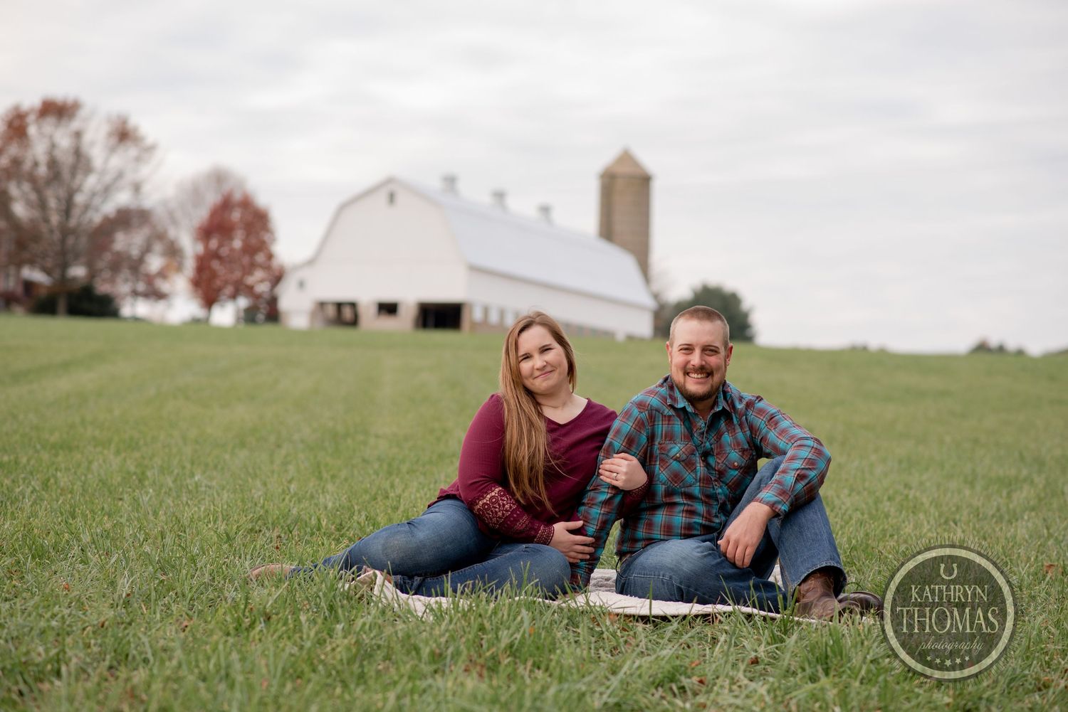 A couple relaxes on a hillside with a classic white barn and silo in the background.