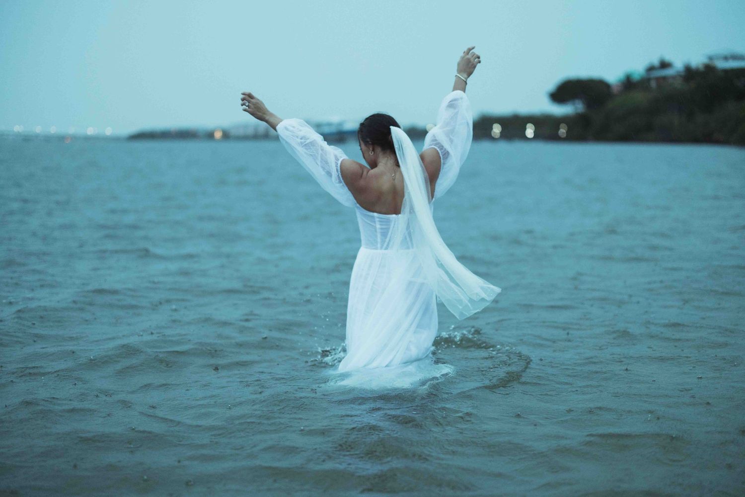 A person in a white dress celebrates with arms raised while standing in ocean water at dusk.