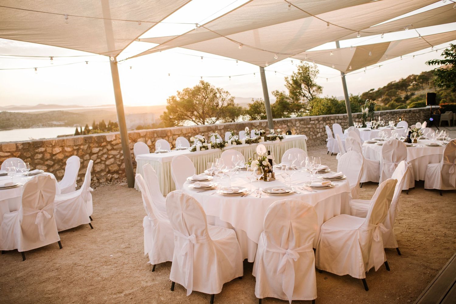 Elegant outdoor wedding reception setup with white-covered tables and chairs under a canopy at sunset.