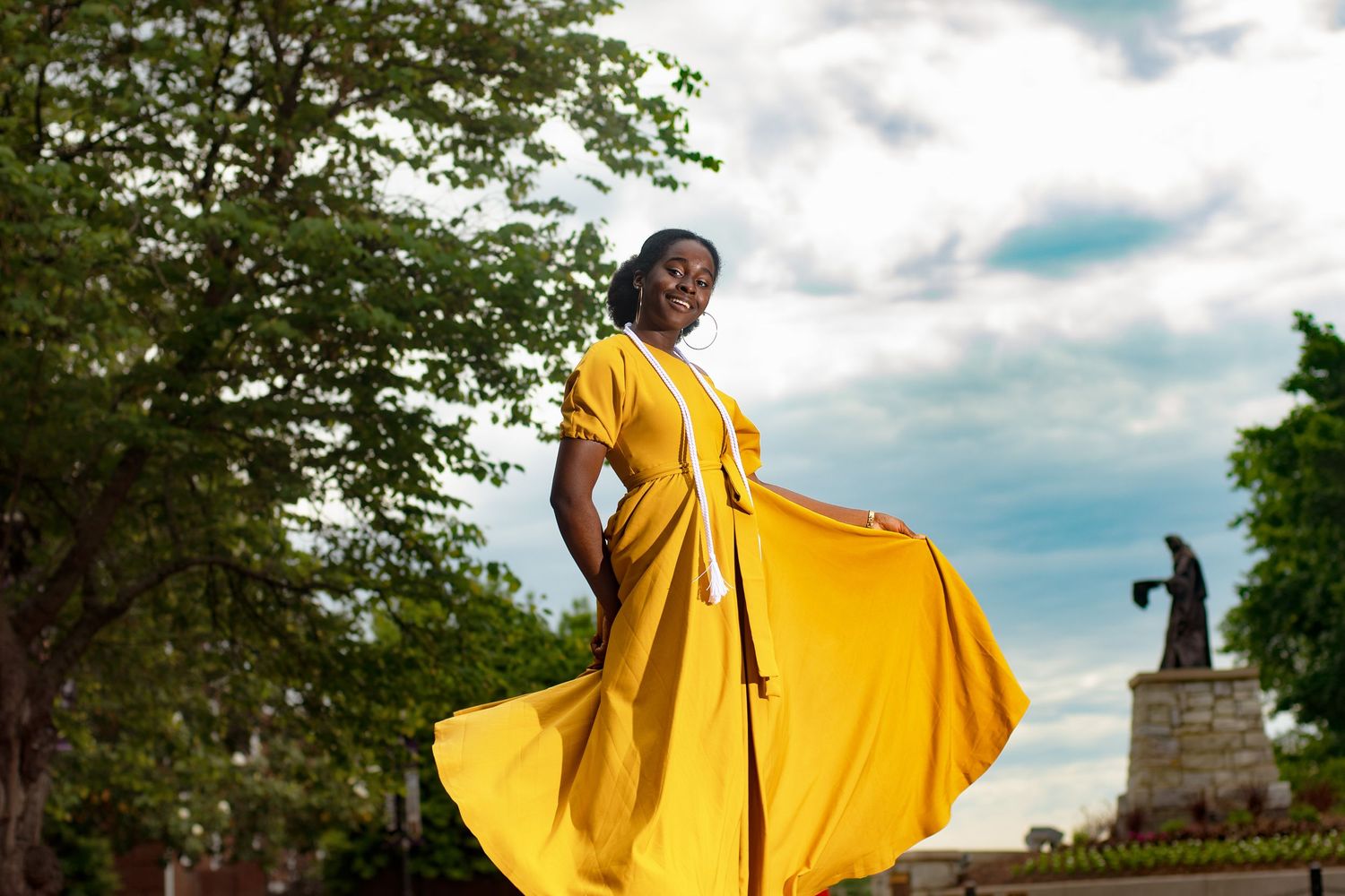 Artistic portrait of person in flowing yellow dress against cloudy sky.